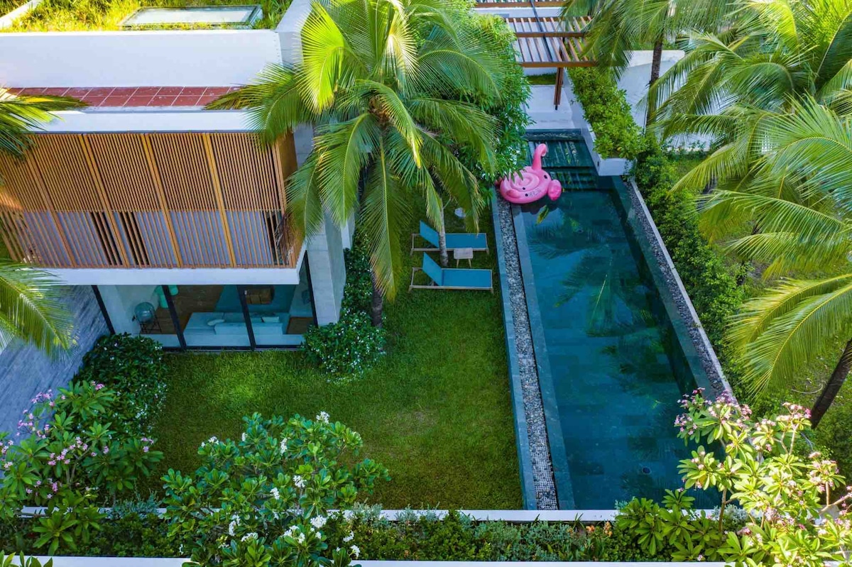 A private pool surrounded by lush tropical greenery is showcased, with sun loungers positioned nearby. A playful pink flamingo float rests on the water's surface, while the villa's modern architecture is visible, featuring wooden slatted detailing and expansive glass windows.