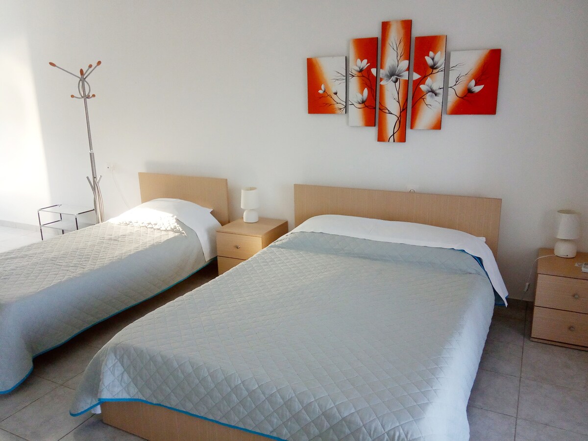 The sleeping area features two beds, one larger and one smaller, with simple bedding in soft hues. A colorful wall art piece showcases flowers against an orange background. Two bedside tables with lamps are positioned beside the beds, enhancing the room's functionality.