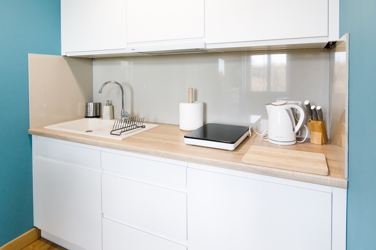 A compact kitchen area features white cabinetry and a light wood countertop. A stainless steel sink is paired with a modern faucet, while kitchen utensils are neatly arranged. An electric kettle and a cutting board are present, with a portable electric burner positioned on the countertop.