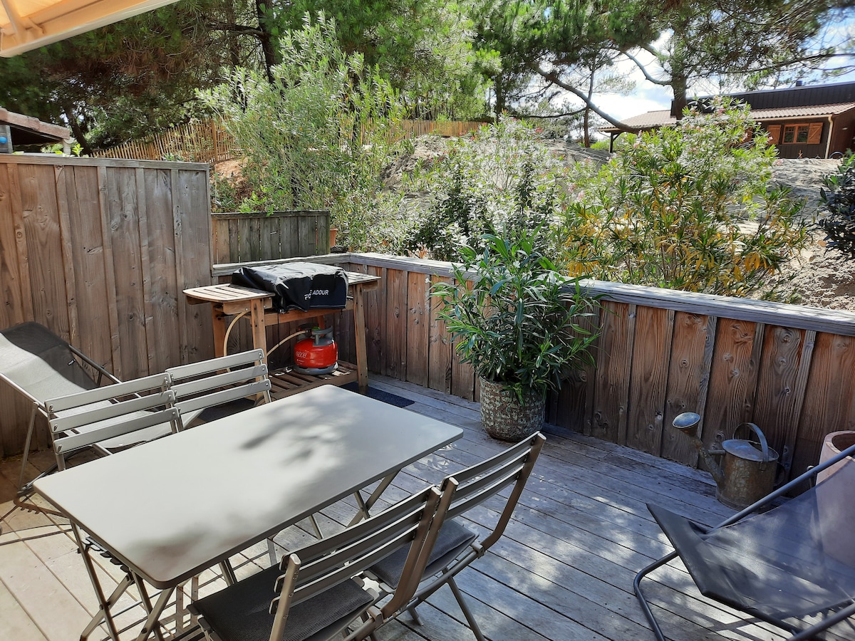 A wooden terrace is featured with a dining table and several folding chairs, surrounded by green plants. In one corner, a charcoal grill is positioned near a storage area. The space is enhanced by natural surroundings, providing a relaxing outdoor setting.