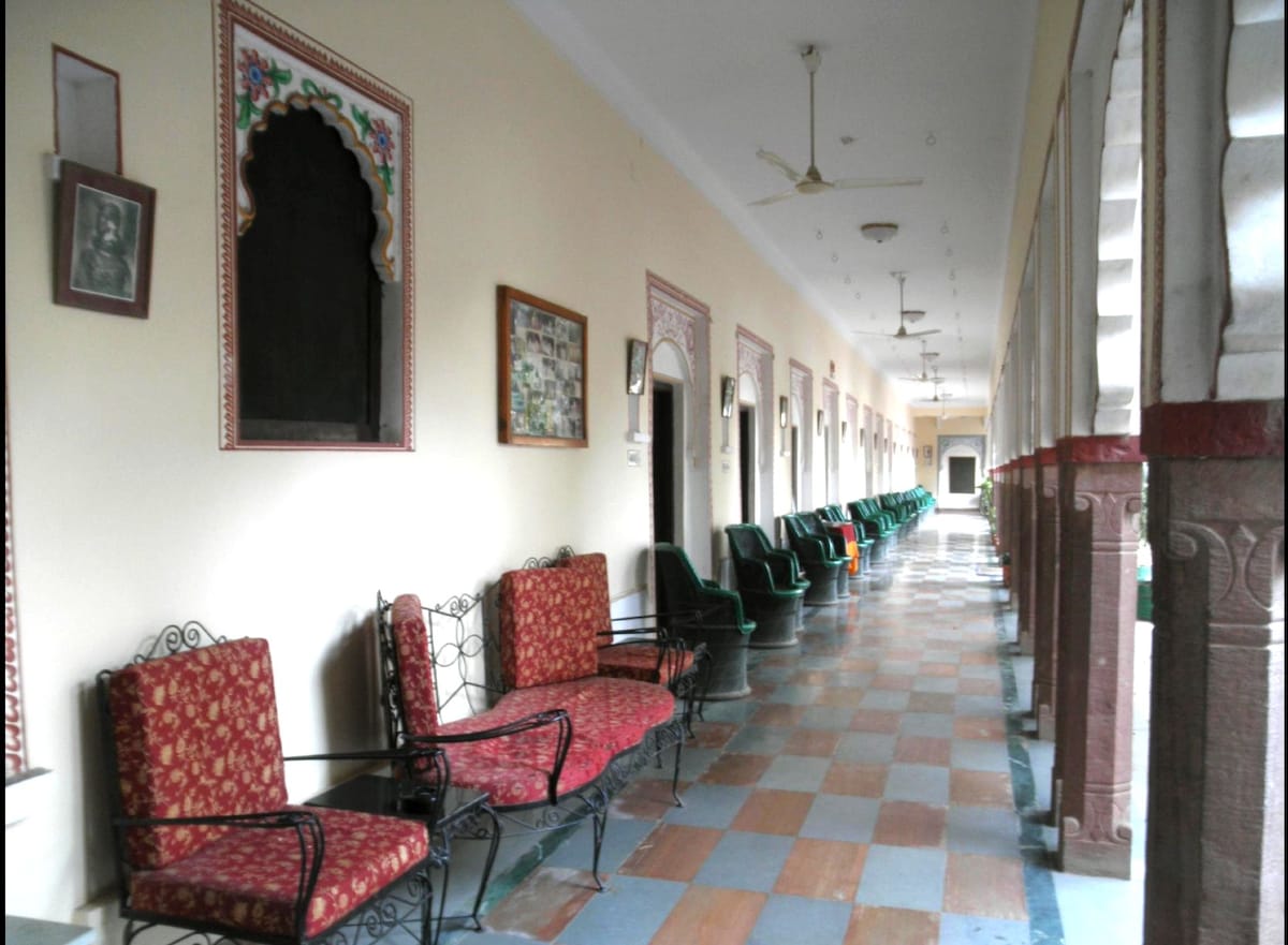 A long corridor features a tiled floor with alternating patterns in blue and terracotta. Ornate arches outline doorways leading to guest rooms. Traditional seating is arranged along the walls, with decorative cushioned chairs in rich red fabric positioned at intervals, inviting relaxation.