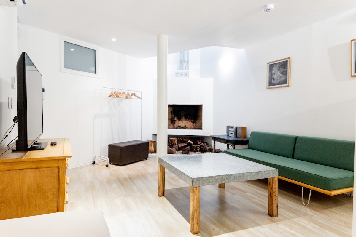 A spacious lower-level living area features a large green sofa and a light grey stone coffee table. A flat-screen TV sits atop a wooden console. A fireplace is visible, surrounded by logs. Large windows provide natural light, and additional seating is arranged nearby.