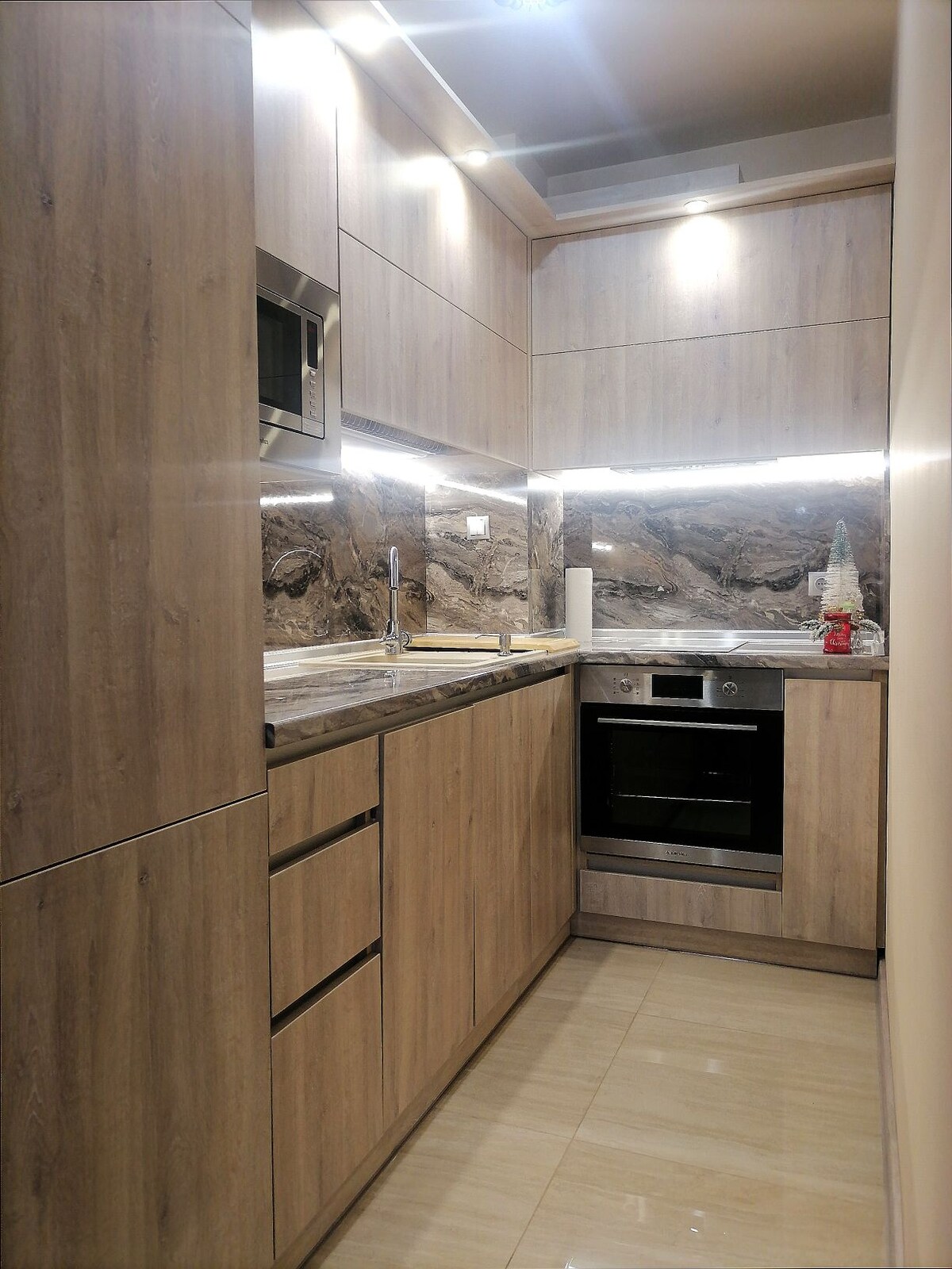 A modern kitchen features wooden cabinetry and sleek countertops. Stainless steel appliances, including a microwave and an oven, are integrated into the design. Bright lighting enhances the clean lines and textures of the space, while a decorative backsplash adds a touch of style.