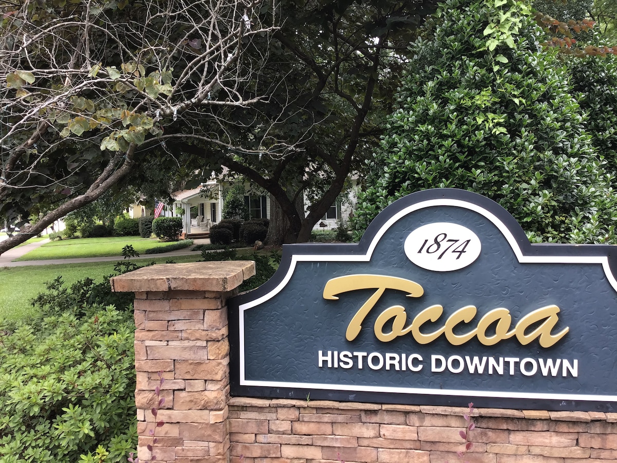The image captures a stone sign welcoming visitors to historic downtown Toccoa. The sign, adorned with the year 1874, is flanked by lush greenery and shaped hedges, providing a glimpse of the tree-lined street and well-maintained properties that characterize the neighborhood.