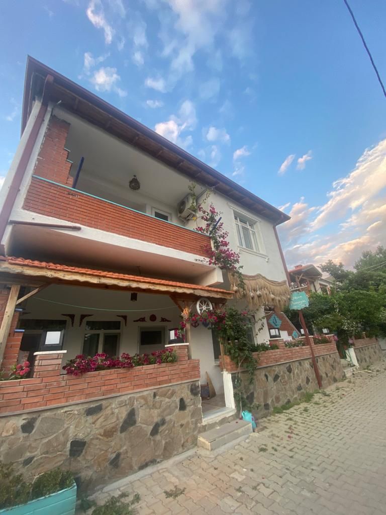 A two-story house is shown with a welcoming façade featuring a combination of brick and stone elements. Flowering plants adorn the balcony and front, while a pathway leads to the entrance. The sky displays soft blue and white clouds, adding to the inviting exterior.