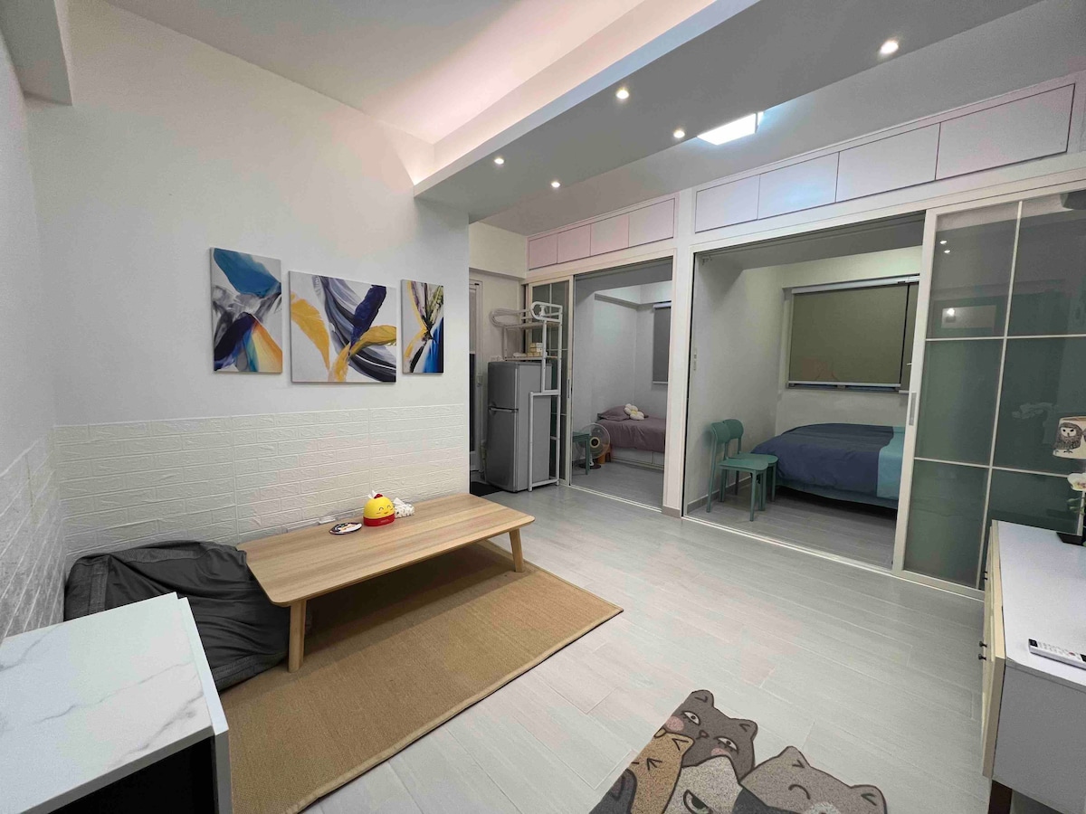 A spacious living area features a low wooden table and a soft rug. Colorful wall art adds visual interest. Glass doors lead to private sleeping areas, and a fridge is visible in the background. A cozy lounge corner is defined by a beanbag chair.