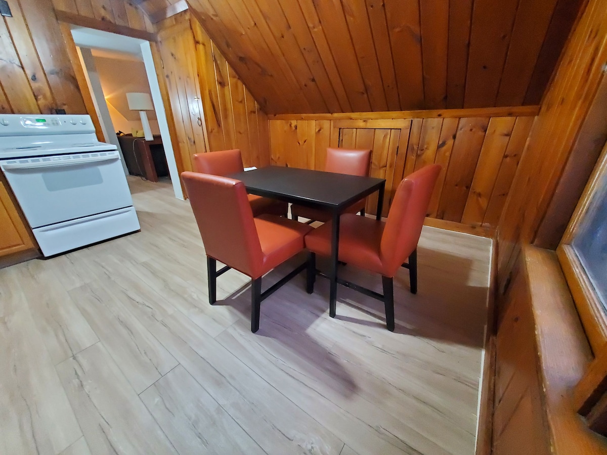 A dining area features a dark rectangular table surrounded by four orange chairs. The wooden walls and ceiling create a rustic ambiance. Soft lighting highlights the warm tones of the space, with a glimpse of the kitchen visible in the background.
