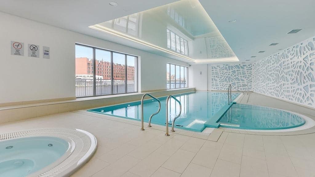 An indoor spa area features a swimming pool with a spacious design, alongside a round jacuzzi. Large windows allow natural light to fill the space, revealing views of the outside. The modern decor includes a decorative wall panel with intricate patterns.