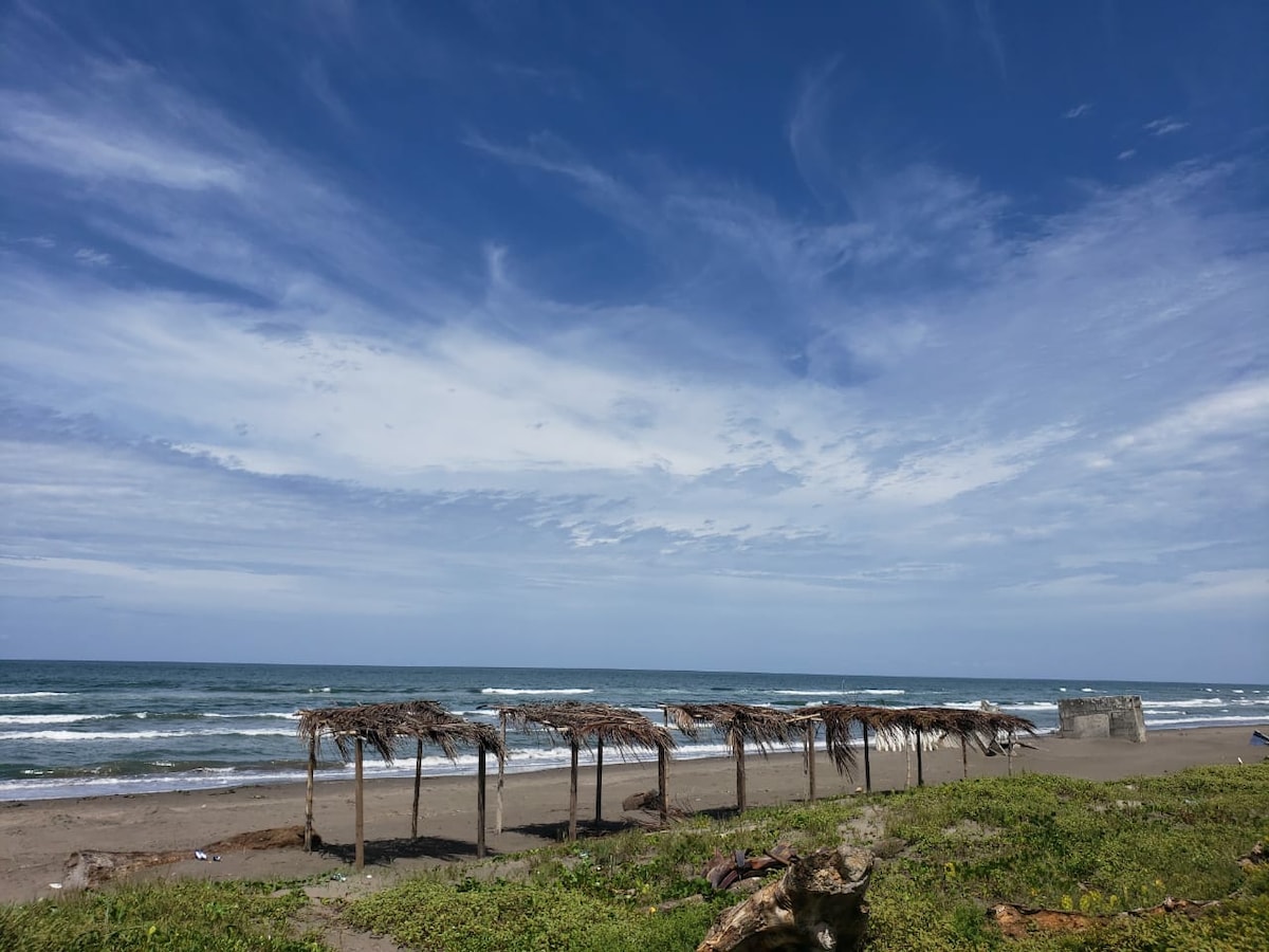 A serene beach scene is depicted, featuring several thatched structures along the shoreline. Gentle waves can be seen lapping at the sandy beach, under a bright sky adorned with wispy clouds. Lush greenery borders the area, enhancing the coastal landscape.