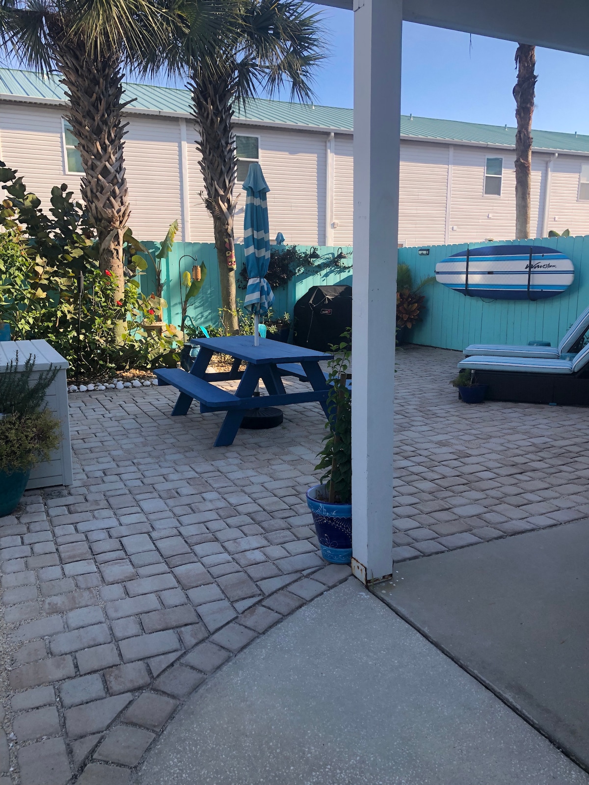 An enclosed courtyard features a picnic table with blue wood, a gas grill, and lounge chairs for relaxation. Surrounding palm trees and colorful plants add greenery to the space. A surfboard is displayed against the turquoise fence, providing a coastal vibe.