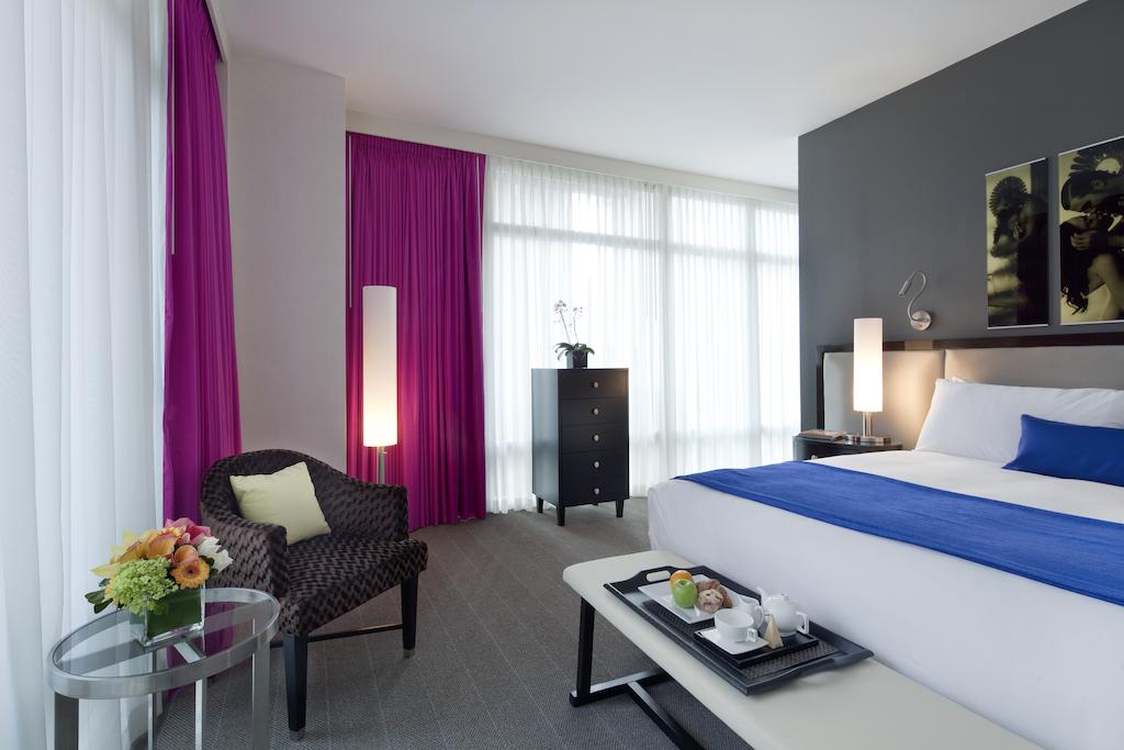 A modern city room features a queen bed with white linens and a blue accent throw. A comfortable chair is positioned beside a glass table, adorned with a floral arrangement and snacks. Sheer curtains allow natural light to fill the space, enhanced by artistic wall decor.