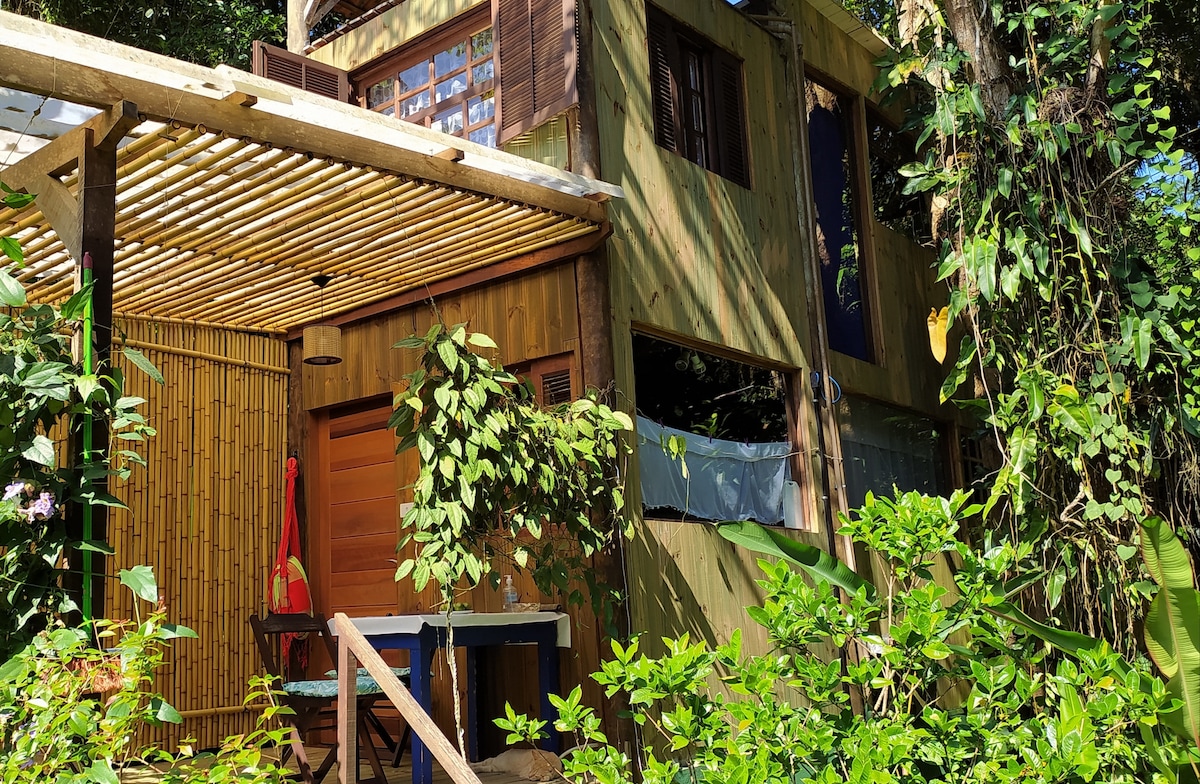 A wooden house is nestled among lush greenery, featuring a shaded patio with wooden furniture. Large windows allow natural light to flow inside, while wooden paneling and vibrant greenery enhance the home’s connection to nature.