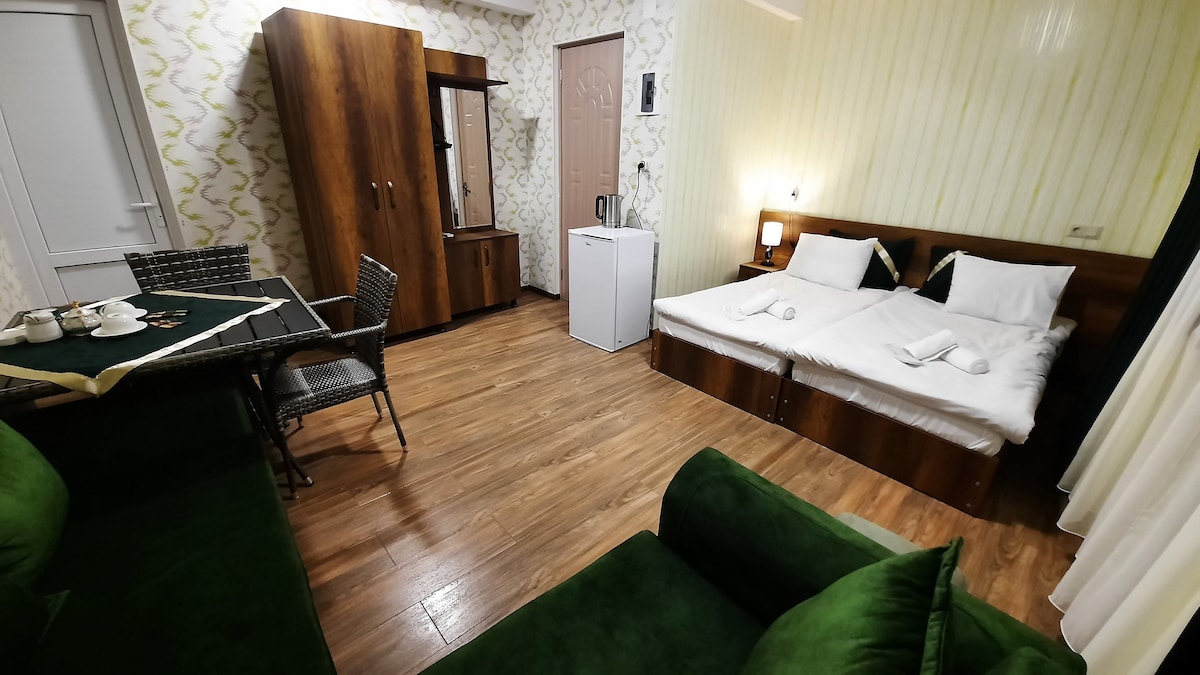 A modern room features a spacious bed setup with light-colored bedding, complemented by a wooden wardrobe. A small table is arranged with diningware, while a refrigerator is positioned nearby. Comfortable seating is provided by a green couch, with warm wooden flooring adding to the overall ambiance.