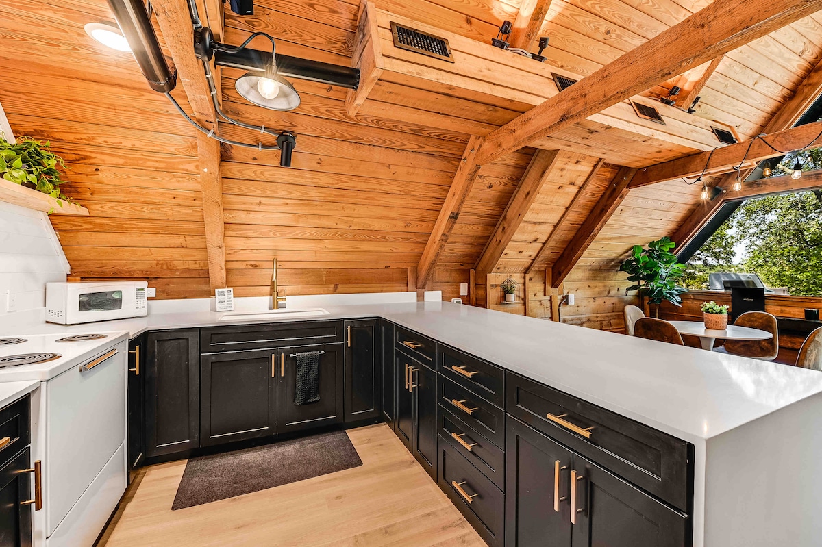 The kitchen space features wooden beams overhead and a light-filled area with ample natural light. Dark cabinetry contrasts with the white countertops, while appliances are neatly arranged for convenience. A dining area with round chairs is visible by a large window, offering a view of surrounding trees.