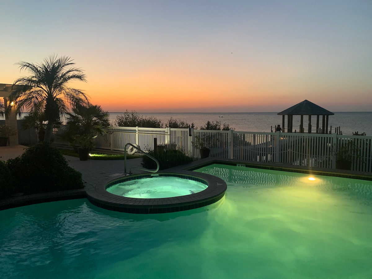 The image captures a serene view of a heated outdoor pool at dusk, with the water illuminated in a gentle glow. In the background, a gazebo is seen against a colorful sunset, while palm trees frame the tranquil scene by the waterfront.
