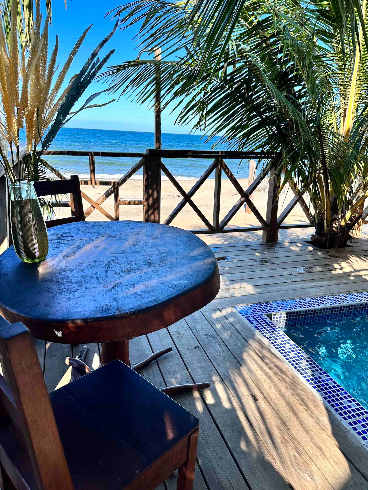 A wooden table and chair are positioned on a deck near a private pool, with lush palm foliage framing the scene. The ocean is visible in the background, enhancing the serene beachside atmosphere.