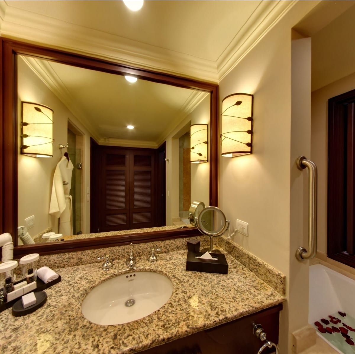 A well-lit bathroom features a granite countertop with a round sink and modern faucets. Soft lighting is provided by stylish sconces on either side of the mirror, enhancing the space's functionality. Folded towels are neatly arranged, and toiletries are organized on the countertop.