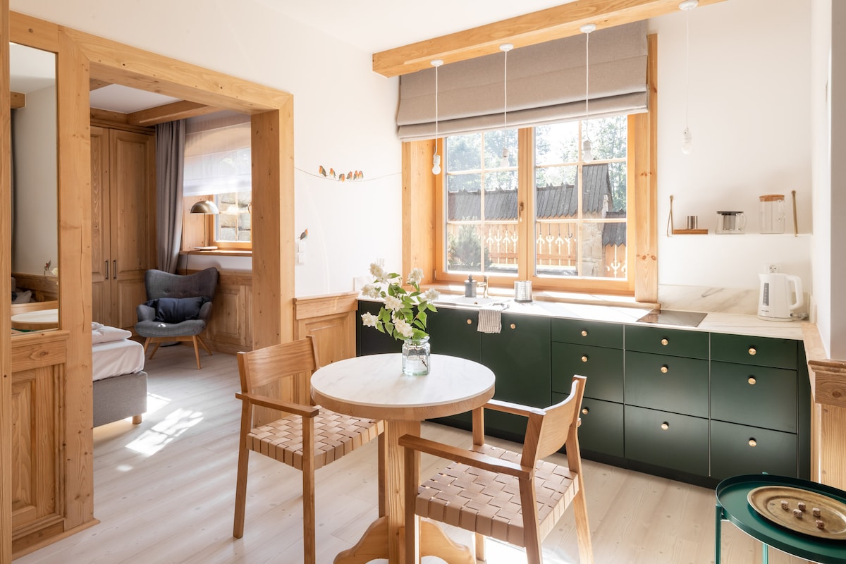 A modern kitchen area features a green cabinetry unit with a small dining table surrounded by two wooden chairs. Natural light fills the space through a large window, showcasing a view of the outdoor surroundings. A cozy seating area is seen in the background.