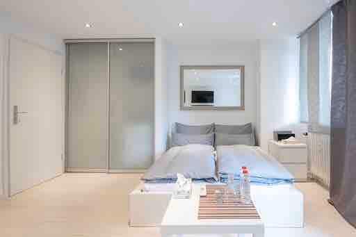 A bright studio apartment showcases a spacious sleeping area with two cozy beds covered in soft bedding. A modern mirrored frame hangs above a small television. Light filters through large windows adorned with light curtains, illuminating the minimalist decor and wooden flooring.