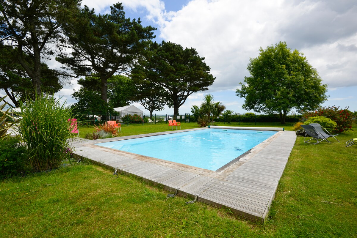 An outdoor heated pool is surrounded by a wooden deck and manicured grass. Lush trees and shrubs provide natural privacy, while lounge chairs are positioned for relaxation. A tent is visible in the background, enhancing the tranquil outdoor setting.