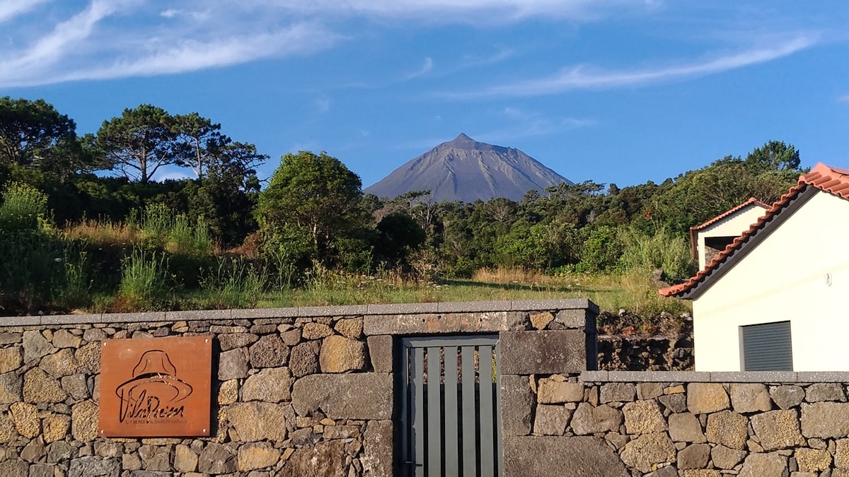 The entrance to Vila Paim is framed by a sturdy stone wall and a wooden gate. A welcoming sign is visible, featuring a logo. The background showcases a prominent mountain under a clear blue sky, with lush greenery on either side, enhancing the natural landscape.