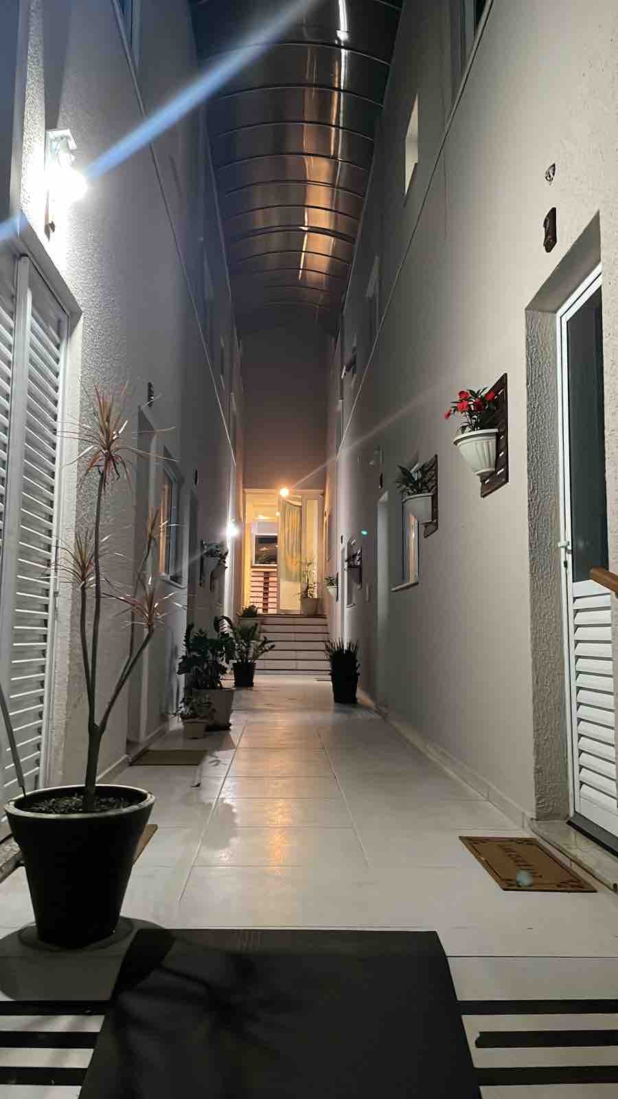 A well-lit corridor is flanked by potted plants and features a clean, tiled floor. The entrance to the duplex is noticeable, with doors on either side leading to separate units. Soft lighting enhances the inviting atmosphere of the space.