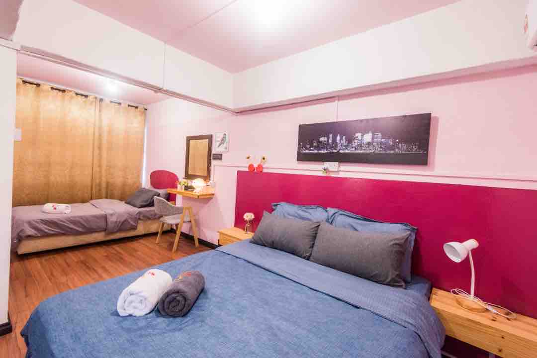 A sleeping area is presented with a spacious bed adorned with gray pillows and a blue coverlet. A single bed is also visible in the background, complemented by a wooden nightstand and a chair. Natural light filters through the golden curtains.