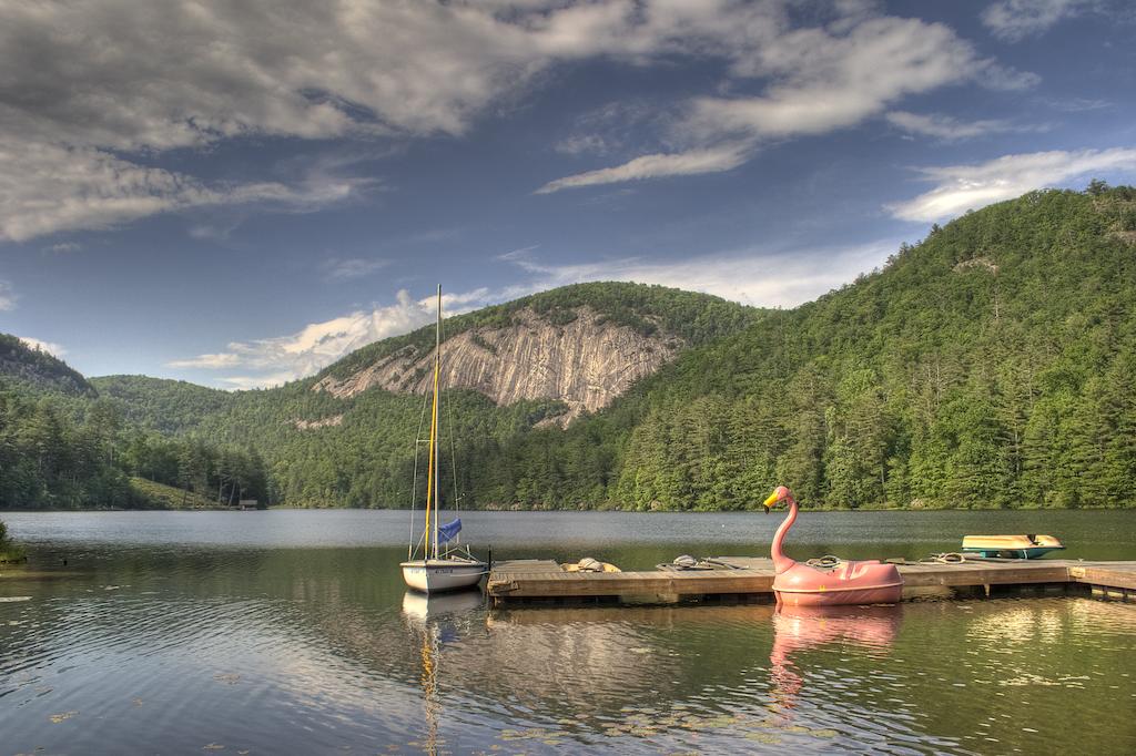 A scenic view of a calm lake surrounded by lush green mountains. A floating dock extends into the water, featuring a pink flamingo pool float and a sailboat moored nearby, with a sail displaying blue and white colors. Dramatic clouds enhance the tranquil atmosphere.