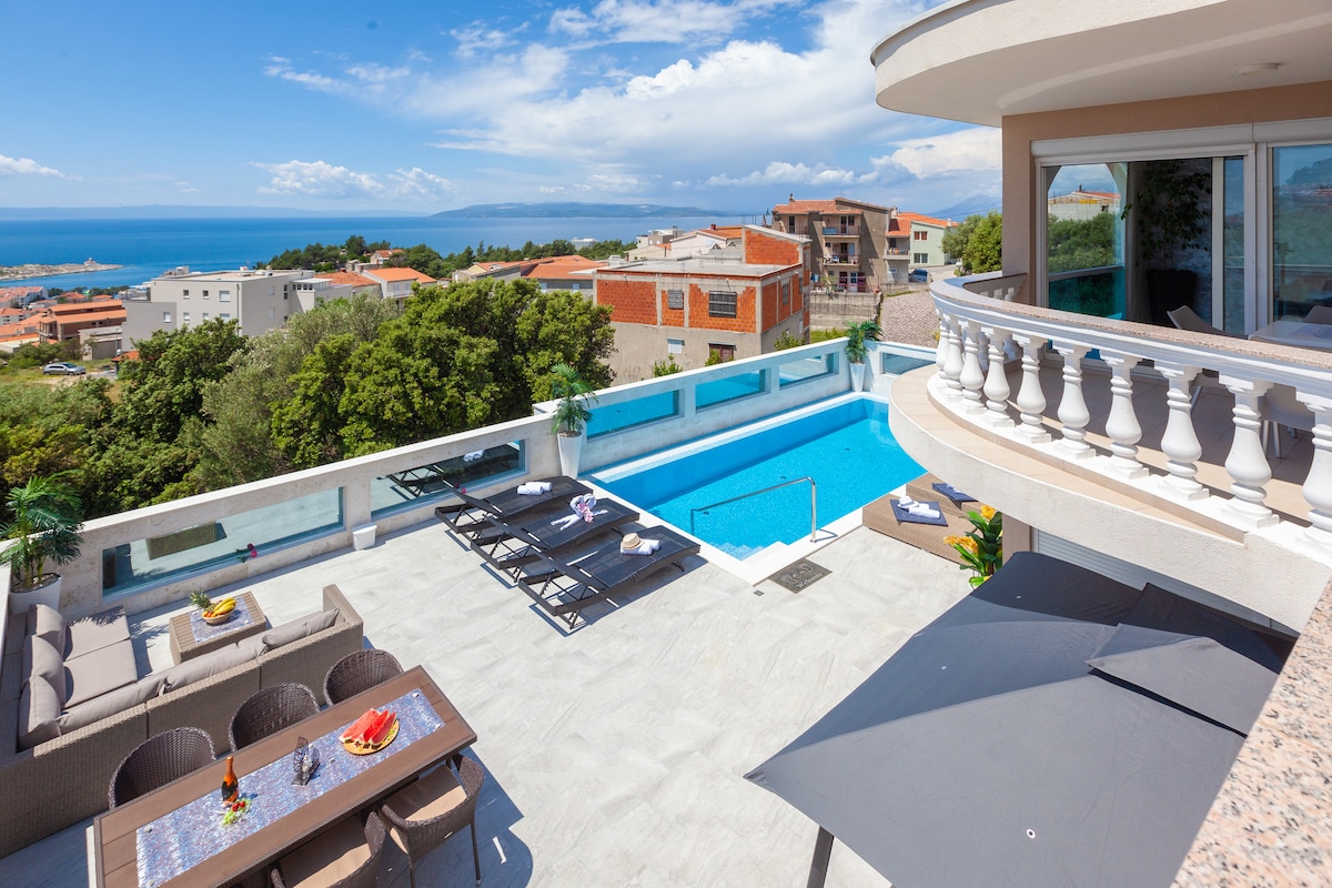 An expansive outdoor terrace features a salt-water pool and several sun loungers arranged for relaxation. A dining table is set up for outdoor meals, with a scenic view of the sea and cityscape in the background, surrounded by greenery.