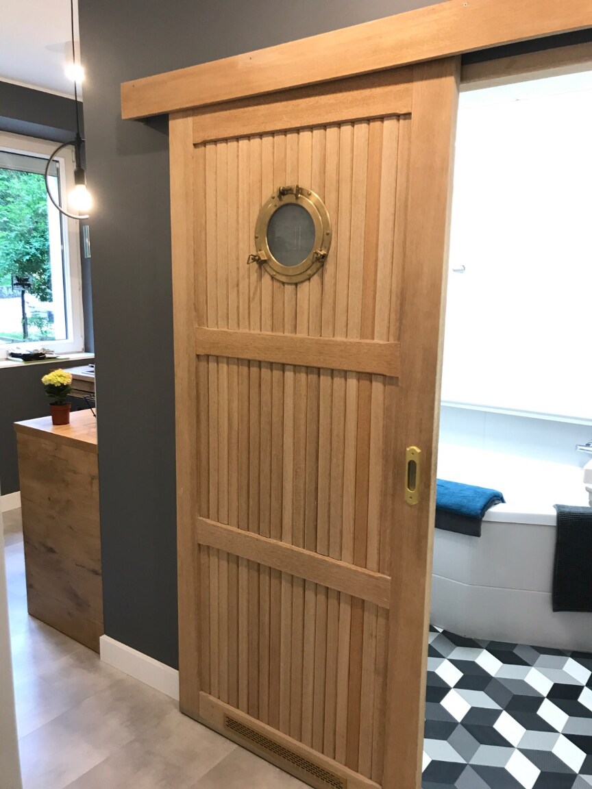 A wooden sliding door features vertical paneling and a circular window with a brass frame. The backdrop includes a modern bathroom partially visible, with a bright color scheme and geometric tiled flooring. A small flower arrangement can be seen on a nearby wooden surface.