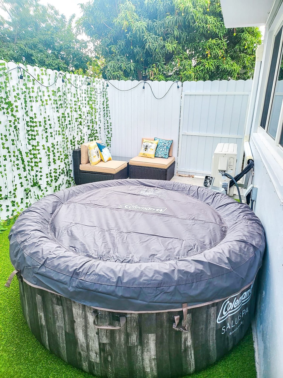 An outdoor space features a hot tub covered with a protective tarp, set against a backdrop of green foliage and a white fence. Comfortable seating is positioned nearby, accented with decorative pillows. The area is designed for privacy and relaxation.
