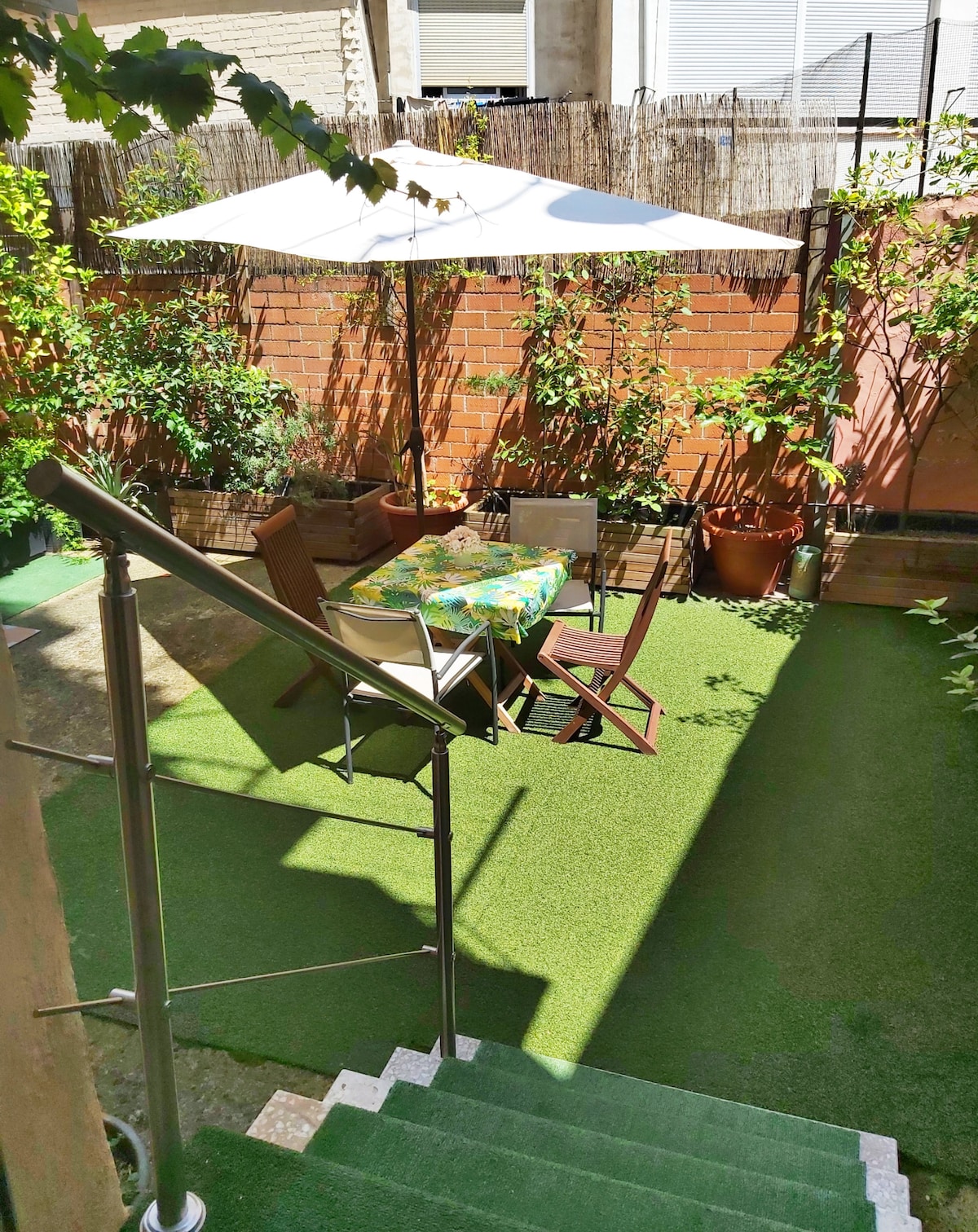 A private courtyard features faux grass and potted plants along the perimeter. A dining table is set with colorful tableware under a large umbrella. Surrounding walls are adorned with greenery, creating a quiet and inviting space for relaxation.