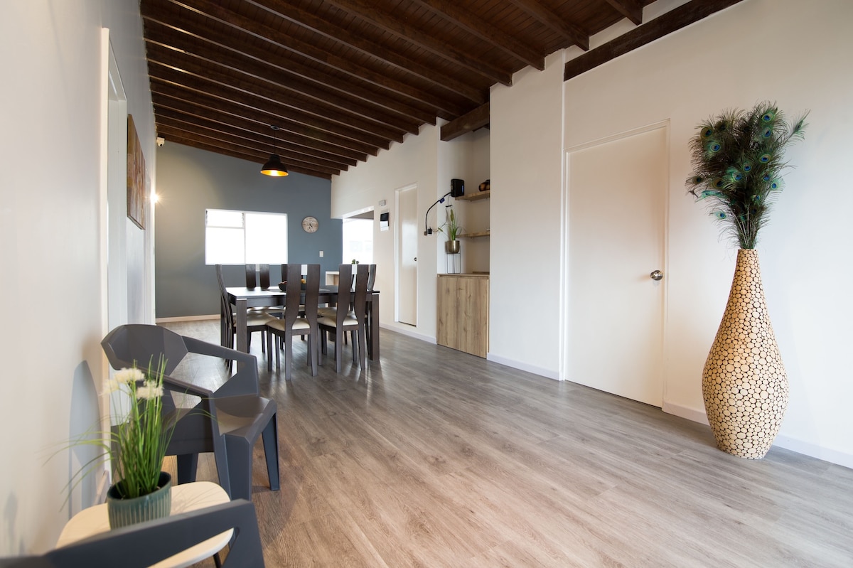 A spacious shared living area features high wooden beams and a contemporary design. A dining table with multiple chairs sits prominently, complemented by stylish seating options in the corner. Natural light streams in through large windows, illuminating the warm wooden floor.
