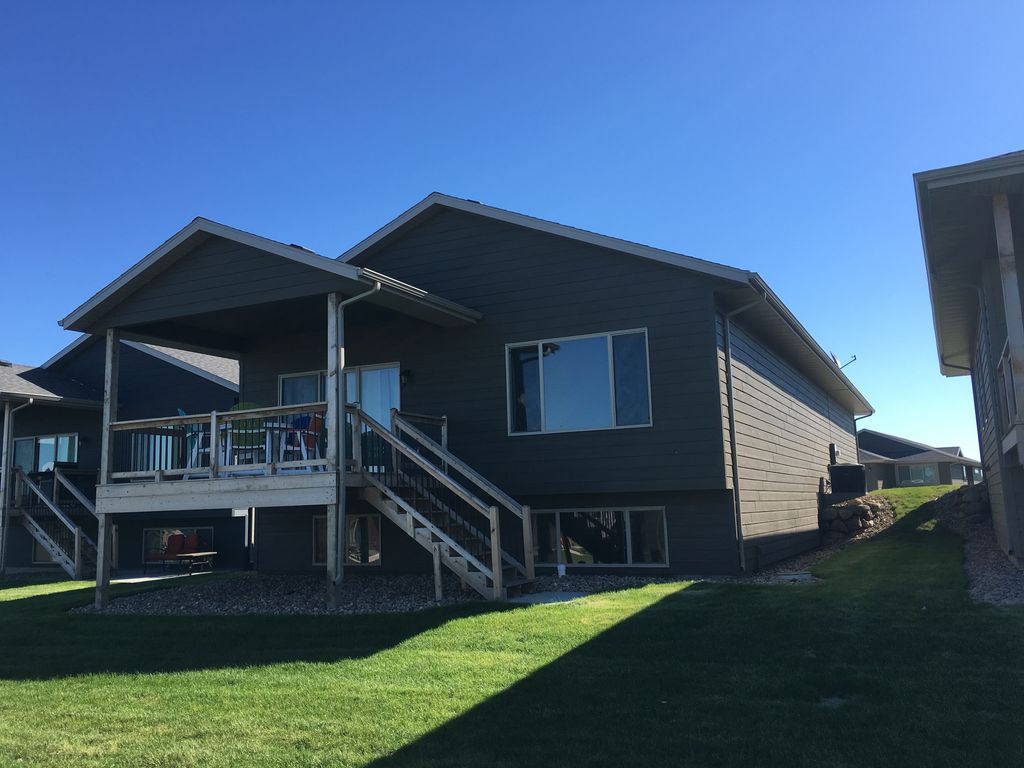 A two-story house is presented, featuring a wide wooden deck that extends from the main floor. Large windows allow for natural light. The exterior is clad in dark siding, with a grassy yard surrounding the property. Clear blue skies are visible above.