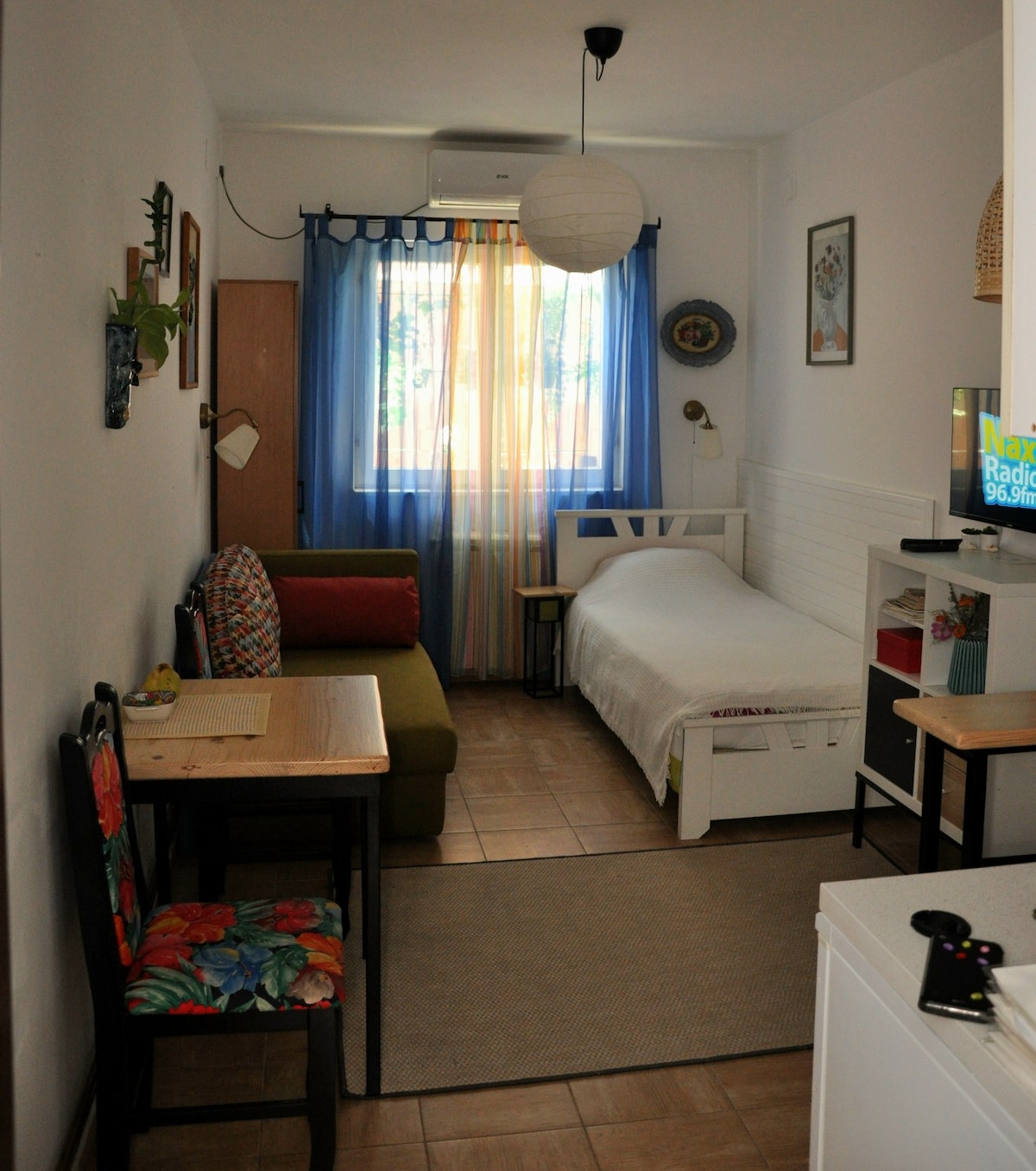 The studio features a bright layout with a spacious bed and a cozy sofa. Natural light floods the room through the colorful curtains. A small table with decorative seating is positioned near the kitchenette, creating a functional area for dining or working.