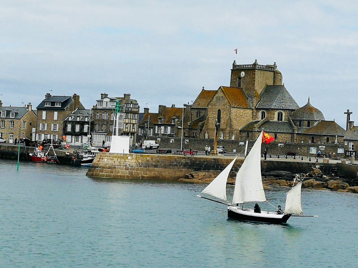 A quaint harbor scene features a traditional sailboat gliding across calm waters, with several charming stone buildings and a prominent church in the background. The coastal structures exhibit various roof styles, while boats are moored along the pier under a soft, overcast sky.