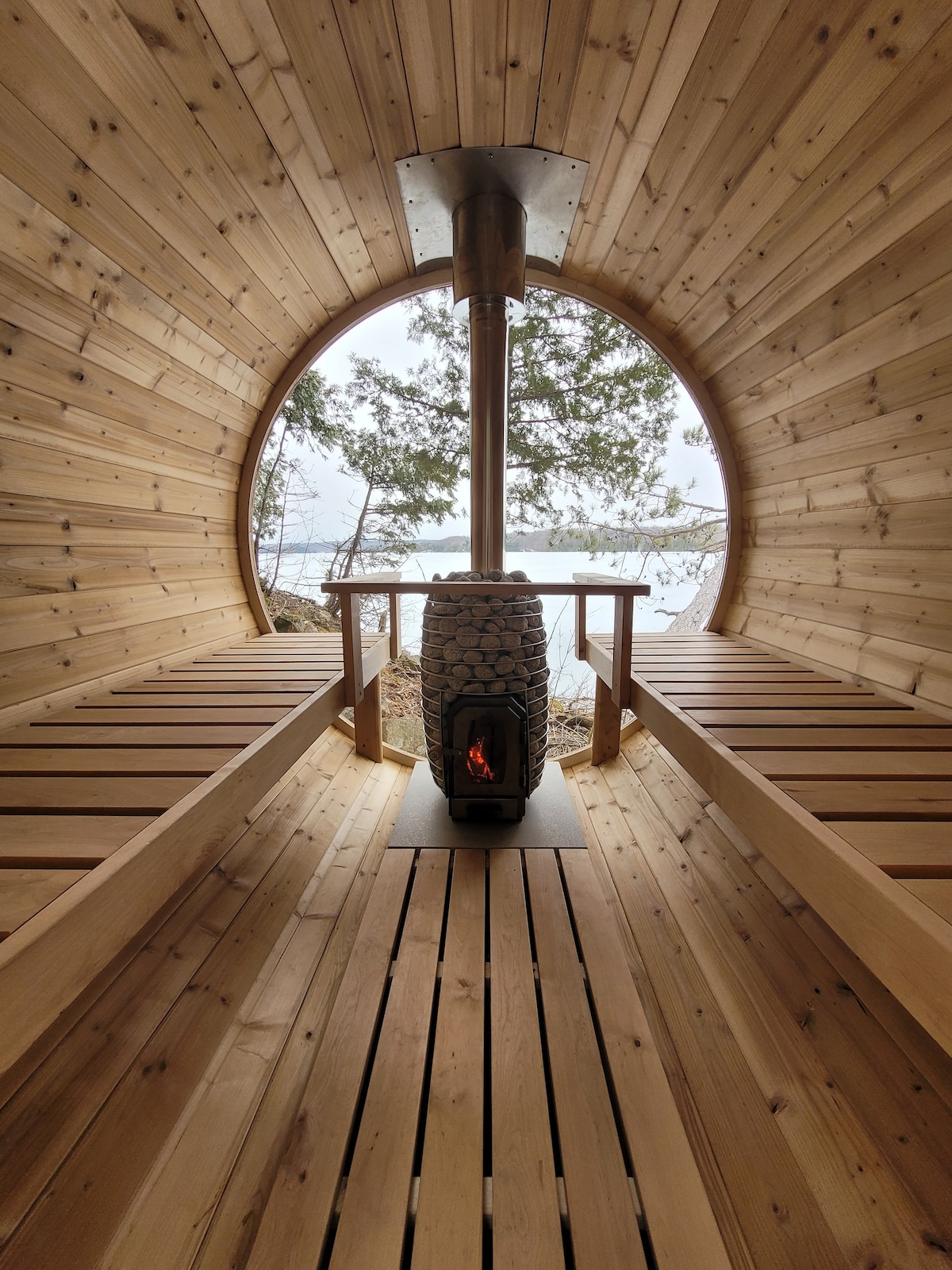 A wood-fired sauna features a rounded interior lined with smooth wooden panels. A centered stone heater radiates warmth, with wooden benches extending along both sides. Large windows offer views of the surrounding trees and lake, creating a serene atmosphere.