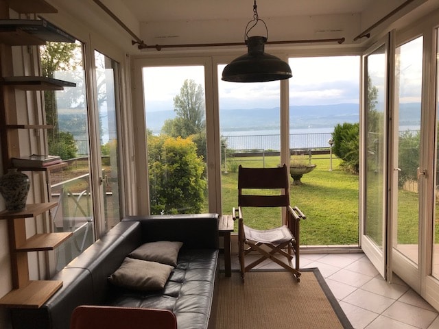 A bright living area features large glass doors that open to a scenic view of the lake. A comfortable black couch is adorned with soft pillows, while a wooden chair complements the space. Lush greenery is visible just outside, enhancing the tranquil atmosphere.