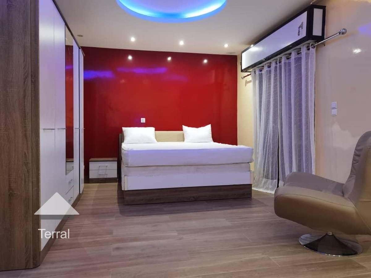 A modern bedroom features a spacious bed with white linens against a bold red wall. Soft lighting is provided by recessed ceiling fixtures, and sheer curtains frame a window. A minimalist wardrobe is positioned to one side, while a comfortable chair is placed nearby.