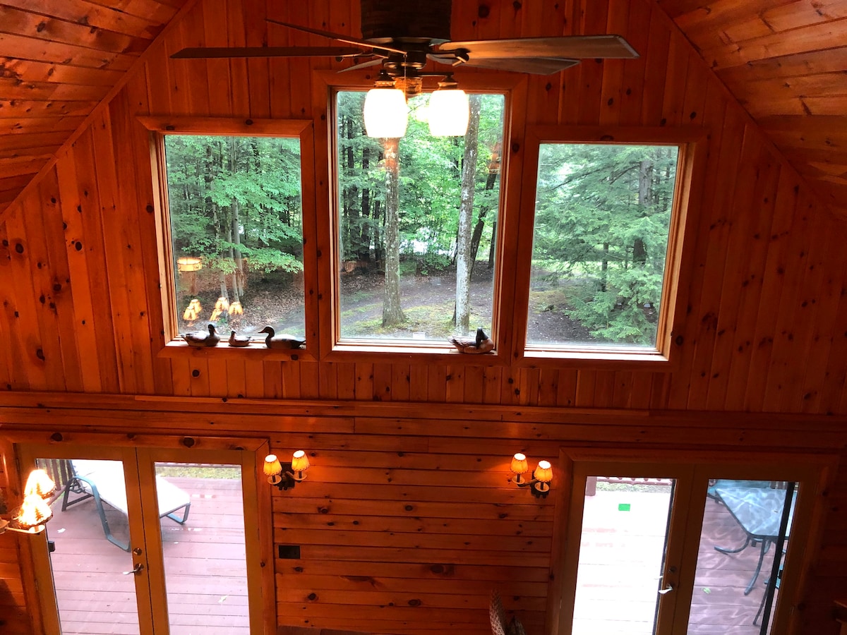 Large windows with unobstructed views of the surrounding trees allow natural light to fill the cabin. The wooden walls and ceiling create a cohesive rustic look, complemented by a ceiling fan and warm lighting fixtures. A balcony area can be seen through the glass doors.