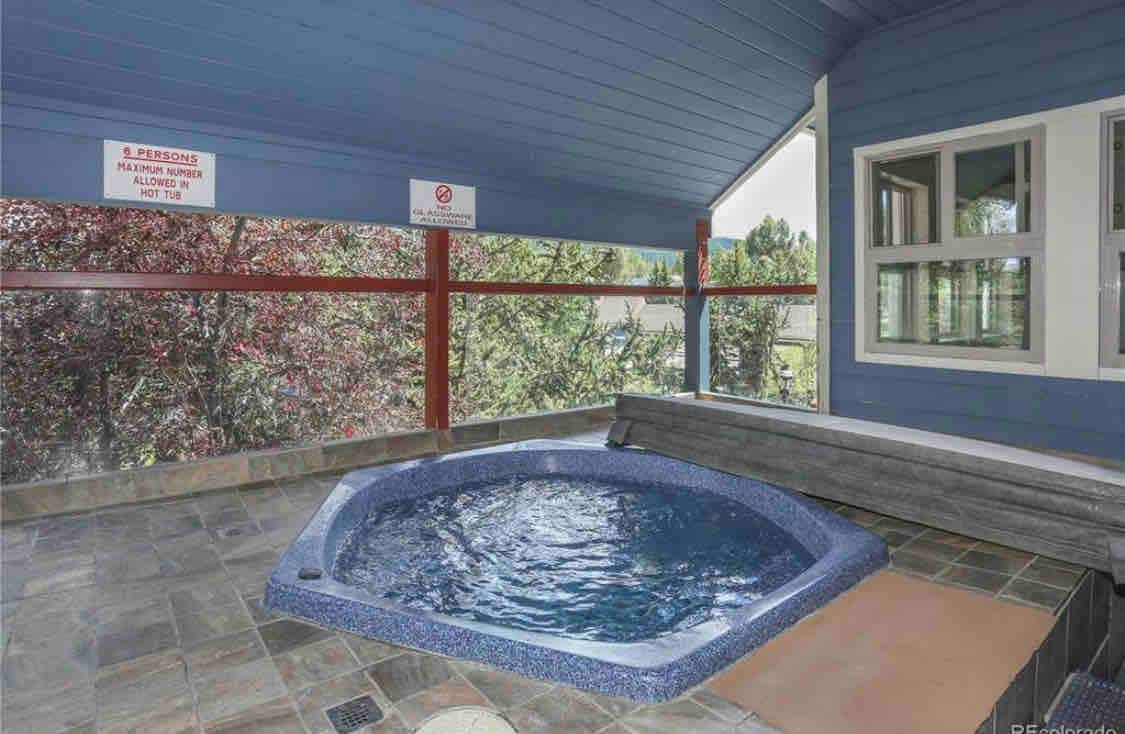 A private hot tub is positioned within a sheltered area, surrounded by large windows that offer views of the nearby greenery. The tub features a rounded shape with a blue interior, complemented by stone flooring that enhances the space's relaxation appeal.
