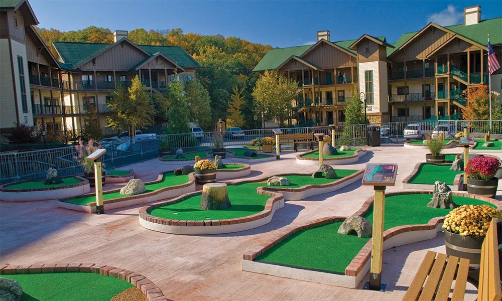 A miniature golf course is set outdoors, featuring creatively arranged green turf with sand traps and decorative rocks. Flowering plants and benches add to the landscape. A backdrop of lodge-style buildings and tree-covered hills enhances the recreational atmosphere.