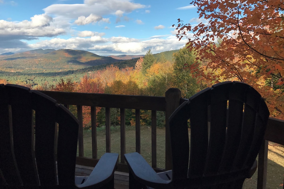 A scenic view is presented from a wooden deck, showcasing rolling mountains and a vibrant tapestry of autumn foliage. Two Adirondack chairs frame the image, inviting relaxation while enjoying the natural beauty that stretches into the distance under a partly cloudy sky.