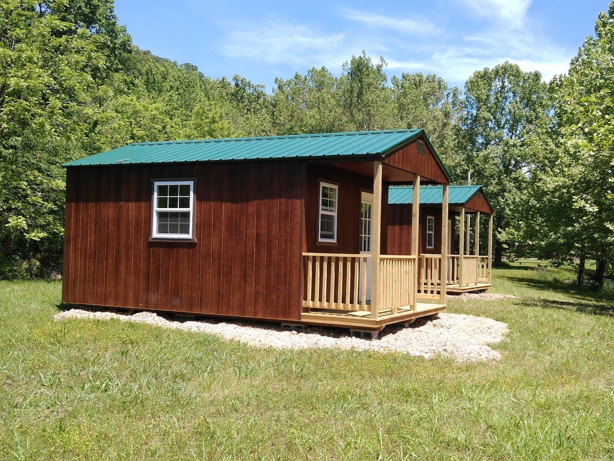 Two wooden cabins are set amidst greenery, featuring a rustic exterior with dark wood siding and metal roofs. Each cabin has a porch with wooden railings. A gravel path leads to the cabins, surrounded by a grassy area and trees in the background.