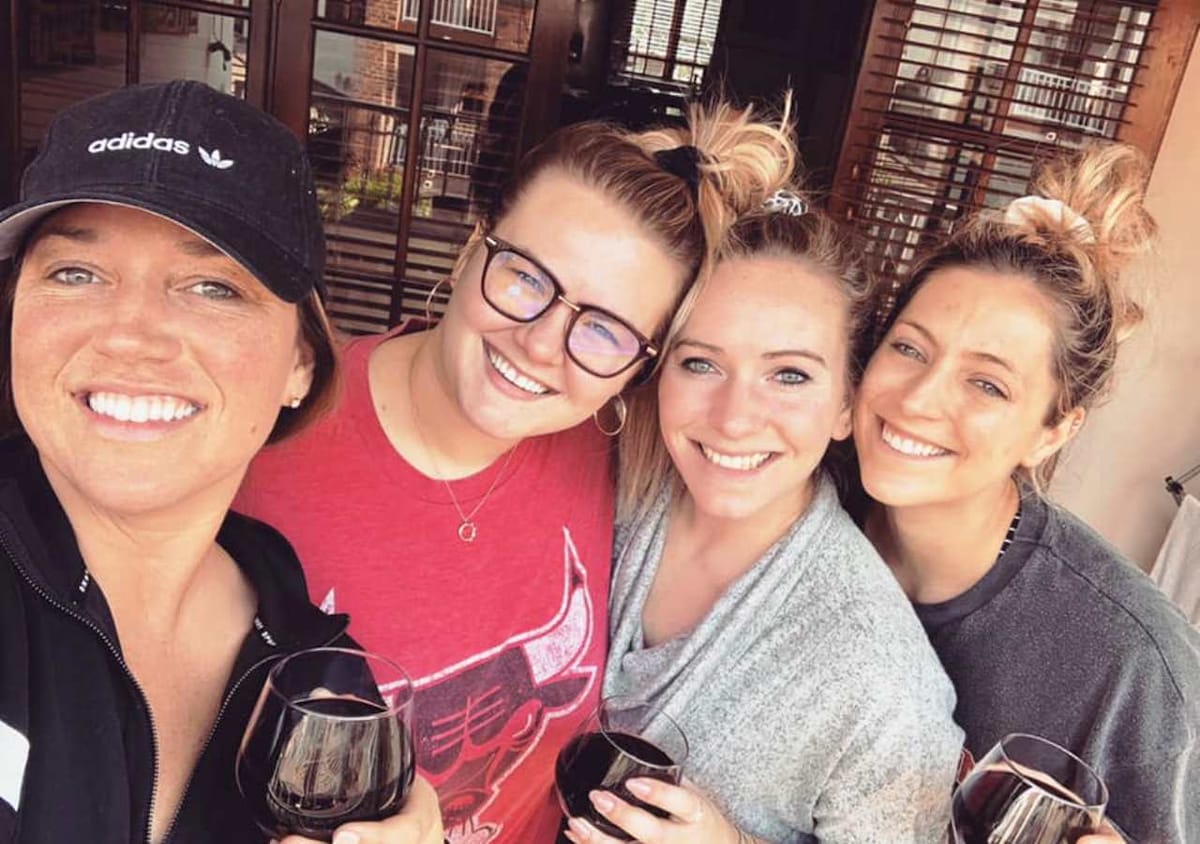 A group of four individuals poses together, each holding a glass of red wine. The warm sunlight filters through wooden blinds in the background, contributing to an inviting atmosphere. Casual attire is worn, with relaxed hairstyles complementing the friendly gathering.