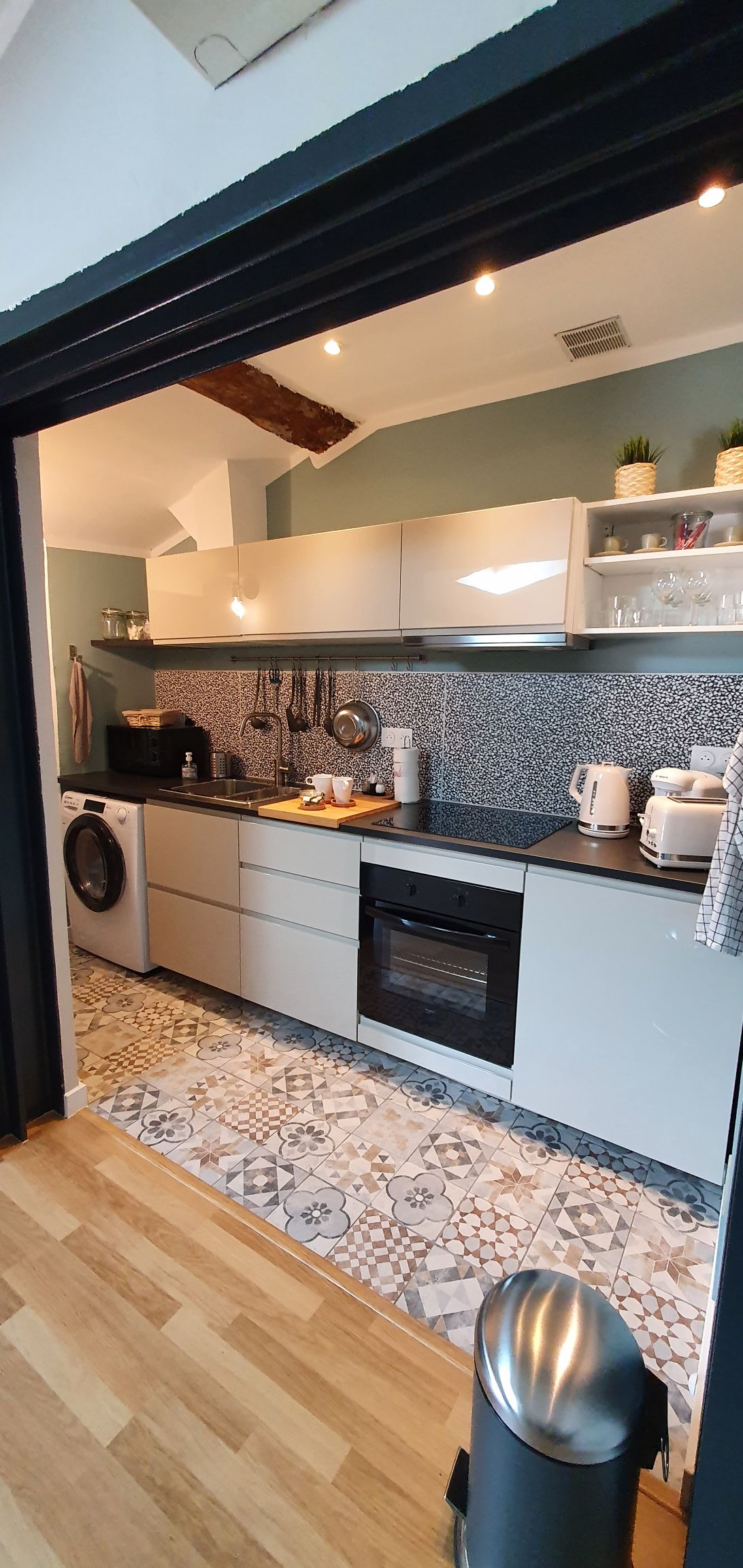 A modern kitchen is equipped with sleek cabinets and a patterned tile floor. Appliances include a refrigerator, washing machine, kettle, and coffee maker, all arranged along a light countertop. Shelves hold various kitchen essentials, promoting a functional cooking space.