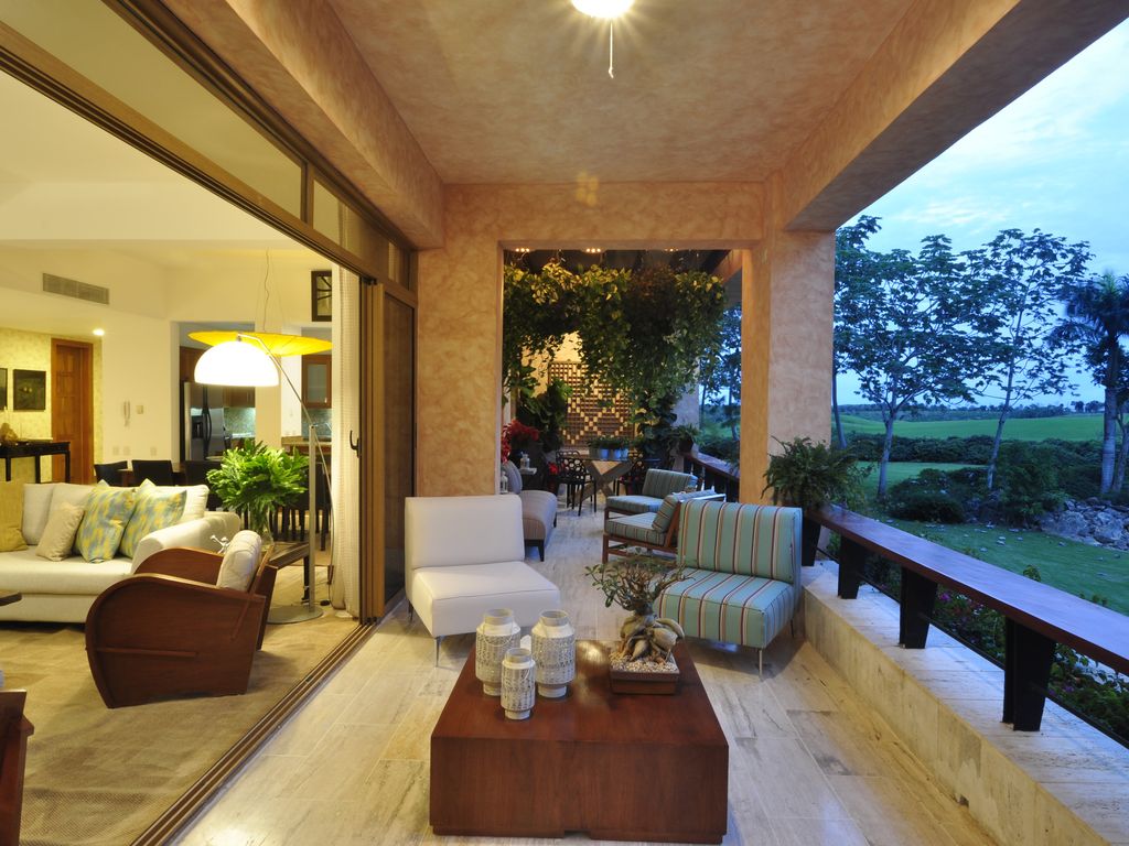 A spacious terrace is furnished with comfortable seating arrangements, including a white sofa and striped chairs. Natural light fills the space, highlighting the marble flooring. Lush greenery and views of the surroundings are visible through expansive glass doors that create an open ambiance.