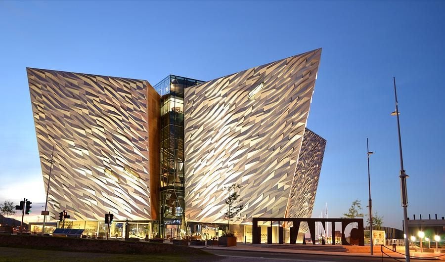 A striking contemporary building featuring a unique, angular façade that reflects the interplay of light. Illuminated at dusk, the surface showcases an array of textured metallic patterns, enhancing its architectural significance. A large 'TITANIC' sign stands prominently in the foreground, contributing to the urban landscape.