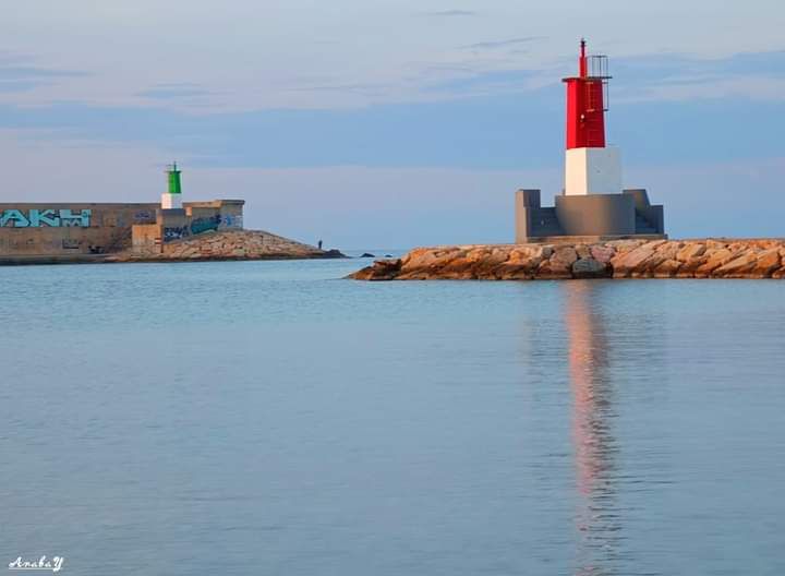 Two lighthouses are positioned along the coastline, one painted in a vibrant red and white design while the other is green. Their reflections can be seen in the calm water, creating a serene coastal scene. The distant horizon features a soft blend of blues and whites.