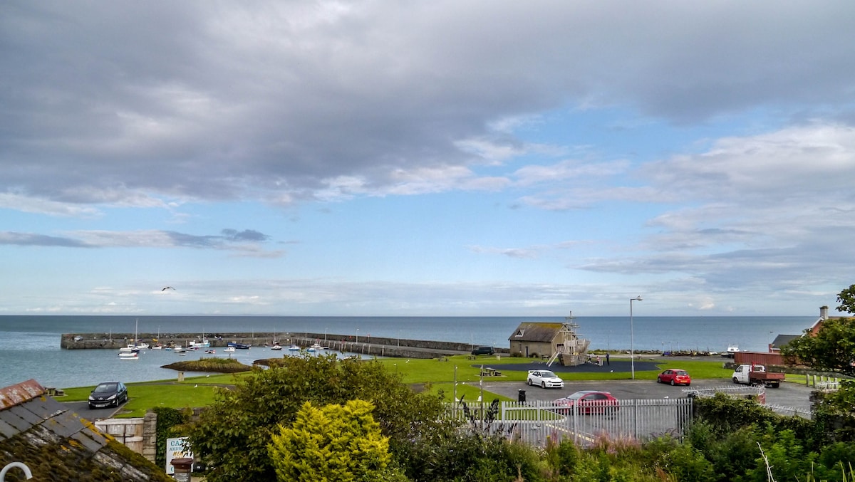 A coastal view captures a serene harbor with multiple boats docked. A small structure is visible near the water, surrounded by grassy areas and parking spaces. The sky features varying shades of grey and blue, reflecting a tranquil atmosphere over the sea.